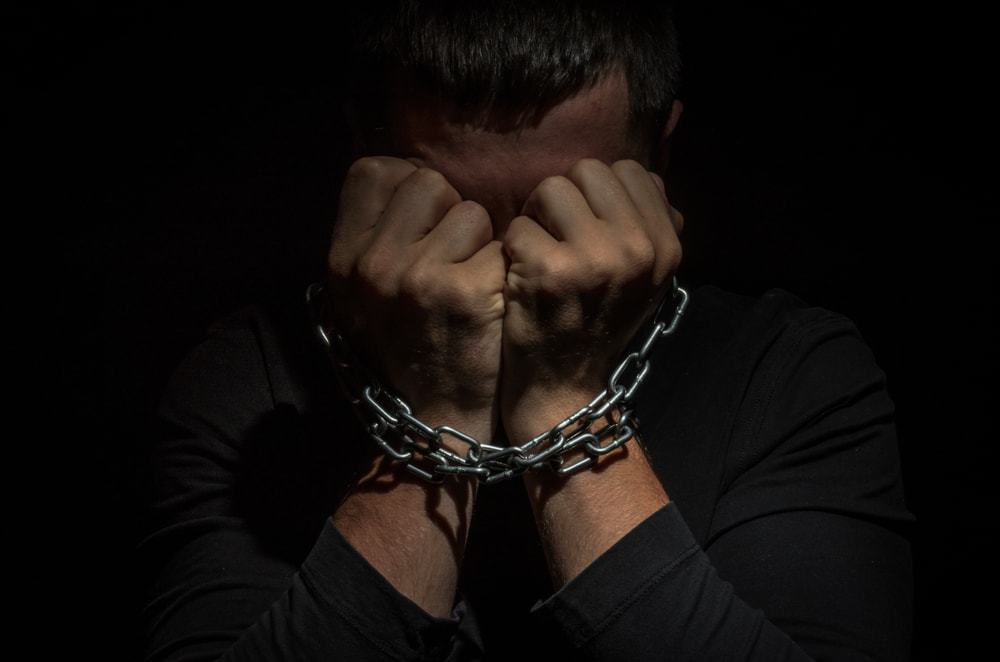 A man in the dark with his hands to his head and a chain around his hands 